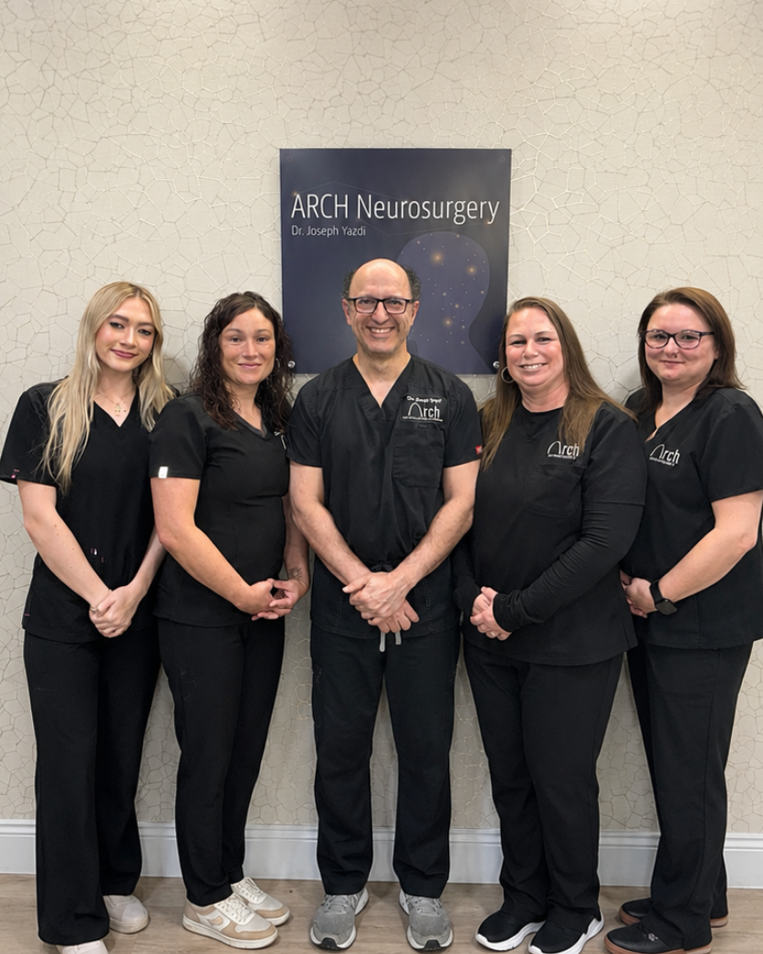Six people, including a man and five women, standing together in front of a beige textured wall with a dark blue 'ARCH Neurosurgery' sign. They are wearing matching black medical scrubs.
