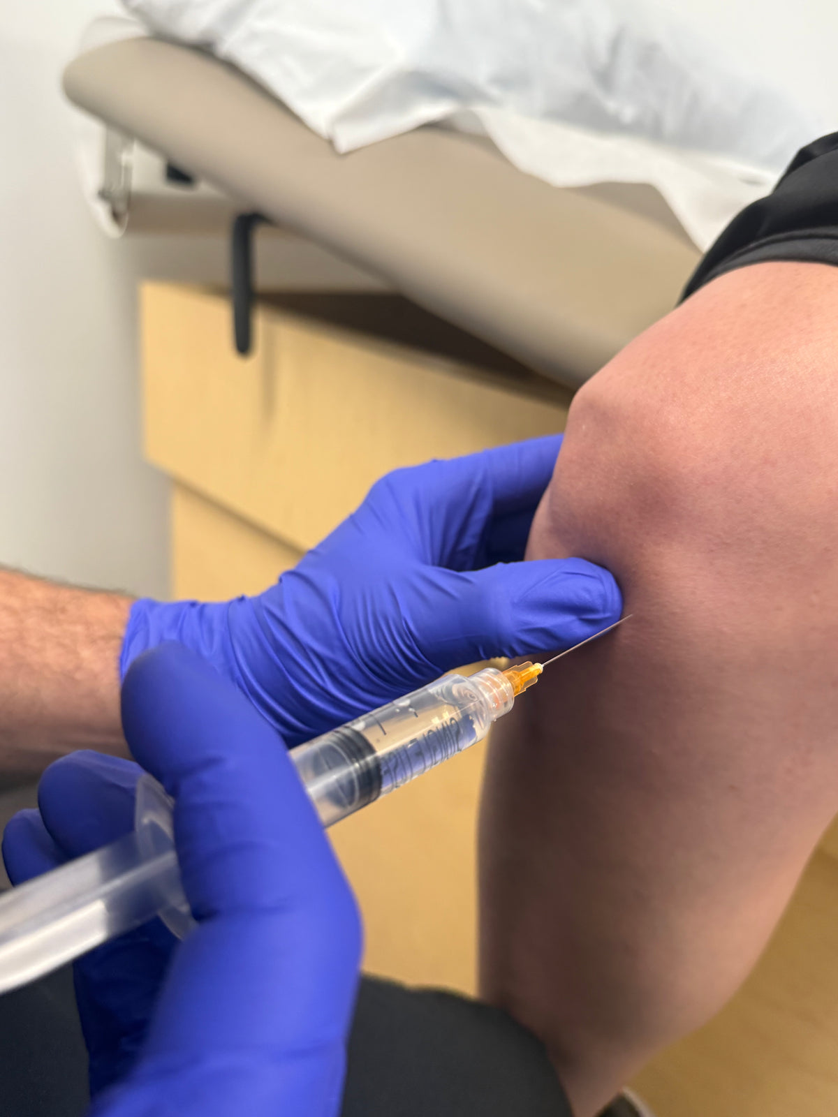 A close-up of a healthcare provider wearing blue gloves administering an injection into a person's knee, with a clinical exam table in the background.