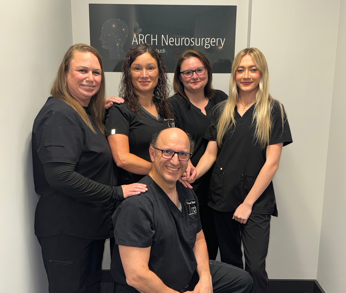 Group of people in black scrubs posing for a photo in front of an ARCH Neurosurgery sign.